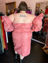 Load image into Gallery viewer, Full-body back view of a size 24 ASOS peachy pink lace sheath dress with a corset-style bodice and oversized ruffle bust and sleeves with puff sleeves on a size 24 model. The photo is taken inside in front of a window, for a combination of natural and overhead lighting.
