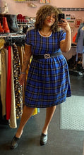 Load image into Gallery viewer, Full-body front view of a size XXL Rudie cobalt blue, white, and black plaid babydoll style midi dress styled with a black belt and black mules on a size 16/18 model.

