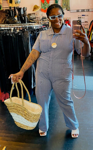 Full-body front view of a size XXXL Nooworks blue and white pinstriped jumpsuit with collar and breast pocket styled with blue sunglasses and an oversize wicker bag on a size 16 model.