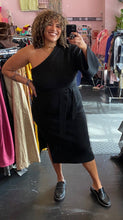 Load image into Gallery viewer, Full-body front view of a size 22/24 Eloquii black one-shoulder bell sleeve sweater dress with tie waist detail and high side slit styled with black loafers on a size 16/18 model.
