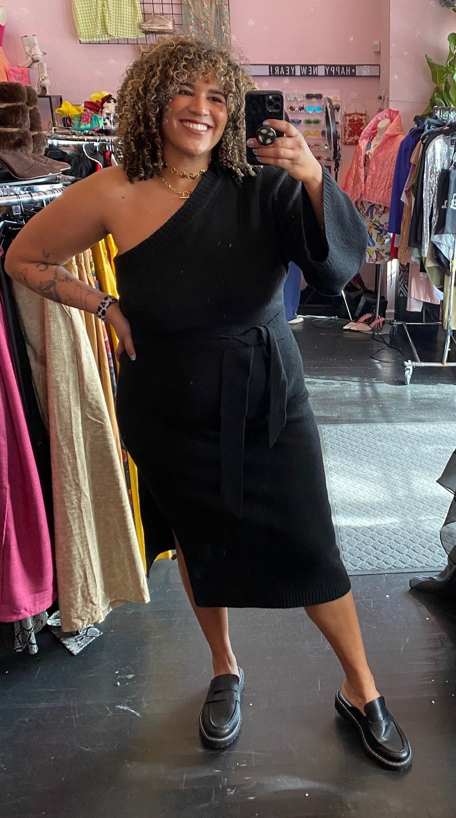 Full-body front view of a size 22/24 Eloquii black one-shoulder bell sleeve sweater dress with tie waist detail and high side slit styled with black loafers on a size 16/18 model.