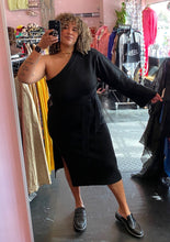 Load image into Gallery viewer, Full-body front view showing off the bell sleeve and side slit of a size 22/24 Eloquii black one-shoulder bell sleeve sweater dress with tie waist detail and high side slit styled with black loafers on a size 16/18 model.
