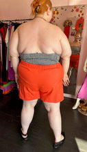 Load image into Gallery viewer, Full-body back view of a pair of size 4X Rebdolls vibrant orange shorts with tortoiseshell button details and pockets styled with a strapless gingham blouse and black pointed flats on a size 22/24 model.
