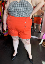 Load image into Gallery viewer, Front view of a pair of size 4X Rebdolls vibrant orange shorts with tortoiseshell button details and pockets styled with a strapless gingham blouse and black pointed flats on a size 22/24 model.
