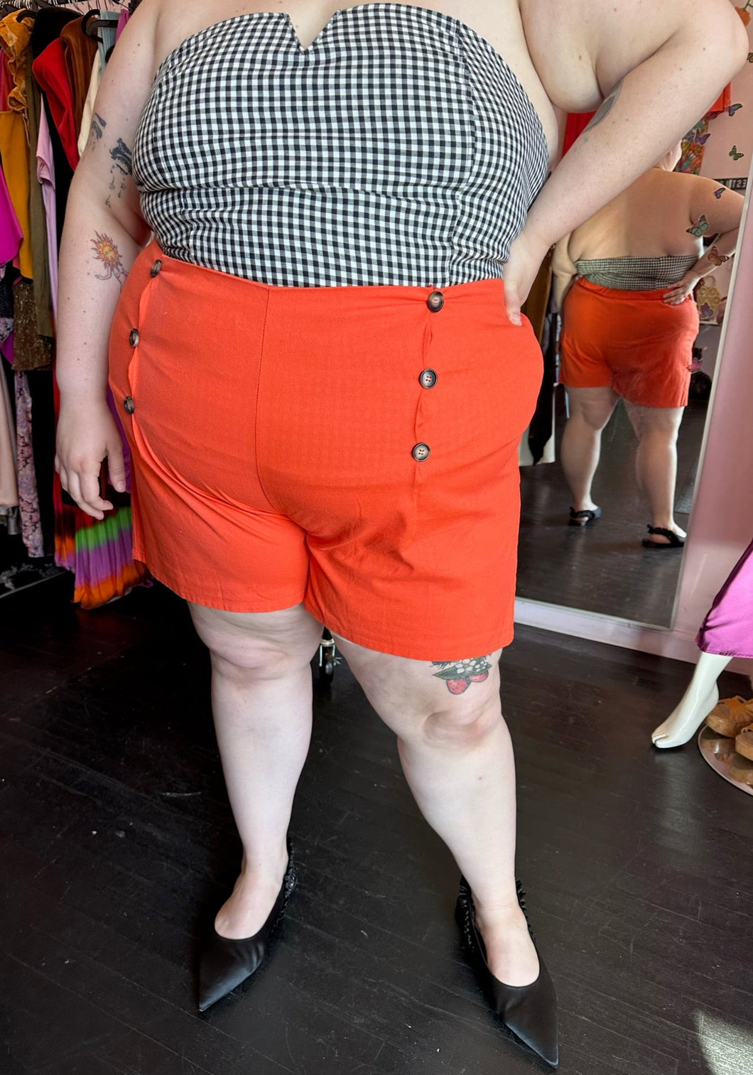 Front view of a pair of size 4X Rebdolls vibrant orange shorts with tortoiseshell button details and pockets styled with a strapless gingham blouse and black pointed flats on a size 22/24 model.