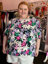 Load image into Gallery viewer, Front view of a size 28 Eloquii black, white, purple, pink, and green geometric floral subtle puff sleeve blouse styled with white shorts on a size 22/24 model.
