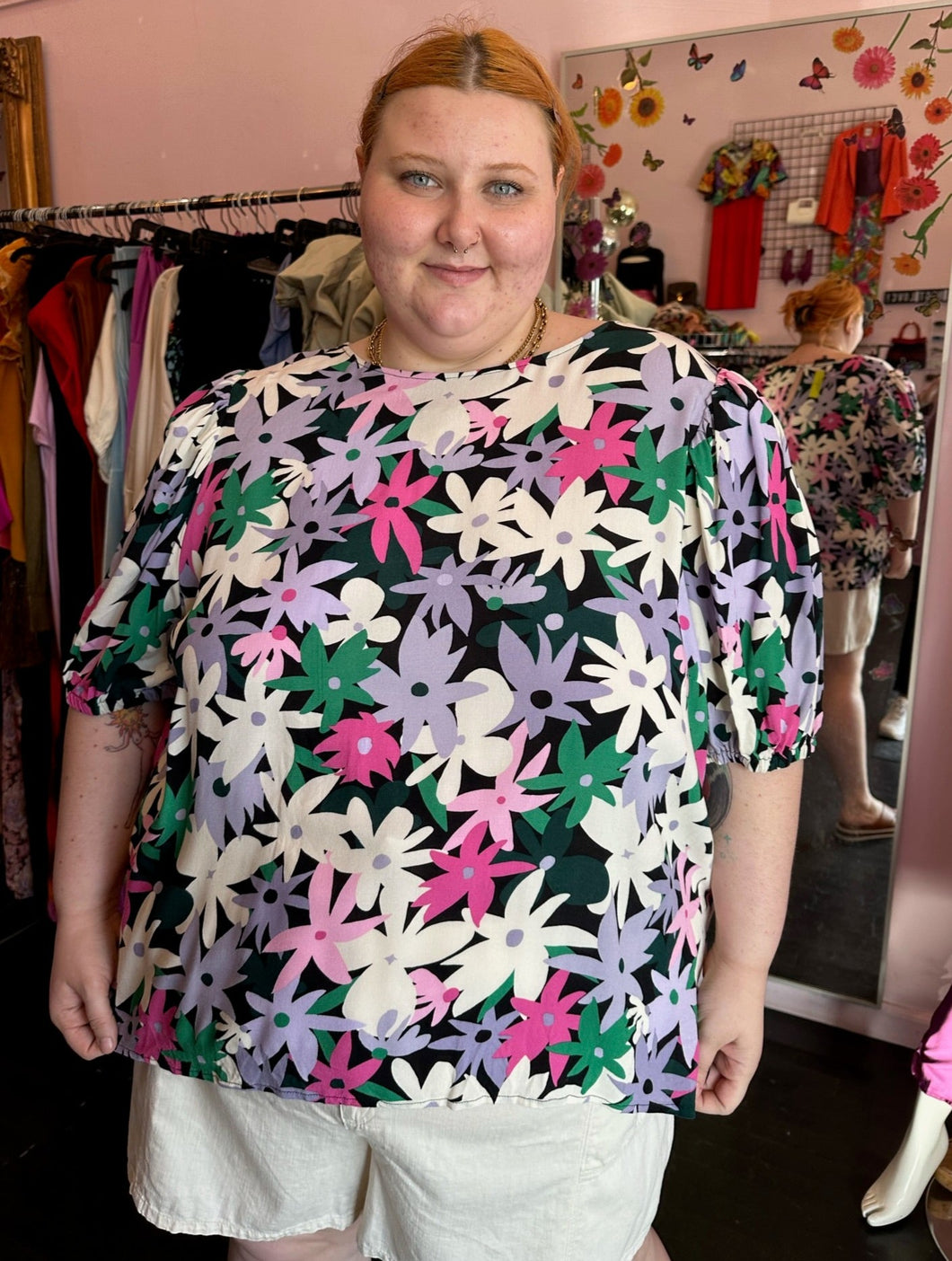 Front view of a size 28 Eloquii black, white, purple, pink, and green geometric floral subtle puff sleeve blouse styled with white shorts on a size 22/24 model.