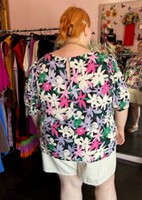 Load image into Gallery viewer, Back view showing off the keyhole detail of a size 28 Eloquii black, white, purple, pink, and green geometric floral subtle puff sleeve blouse styled with white shorts on a size 22/24 model.
