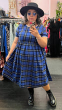 Load image into Gallery viewer, Full-body front view of a size XXL Rudie cobalt blue, white, and black plaid babydoll style midi dress styled with a black hat and black boots on a size 14/16 model.
