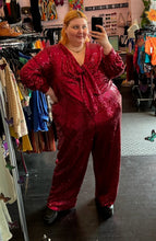 Load image into Gallery viewer, Full-body front view of a size 28 Eloquii hot pink sequin jumpsuit with tie-front bust detail, pockets, and a full lining styled with black boots on a size 22/24 model.
