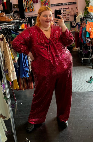 Full-body front view of a size 28 Eloquii hot pink sequin jumpsuit with tie-front bust detail, pockets, and a full lining styled with black boots on a size 22/24 model.