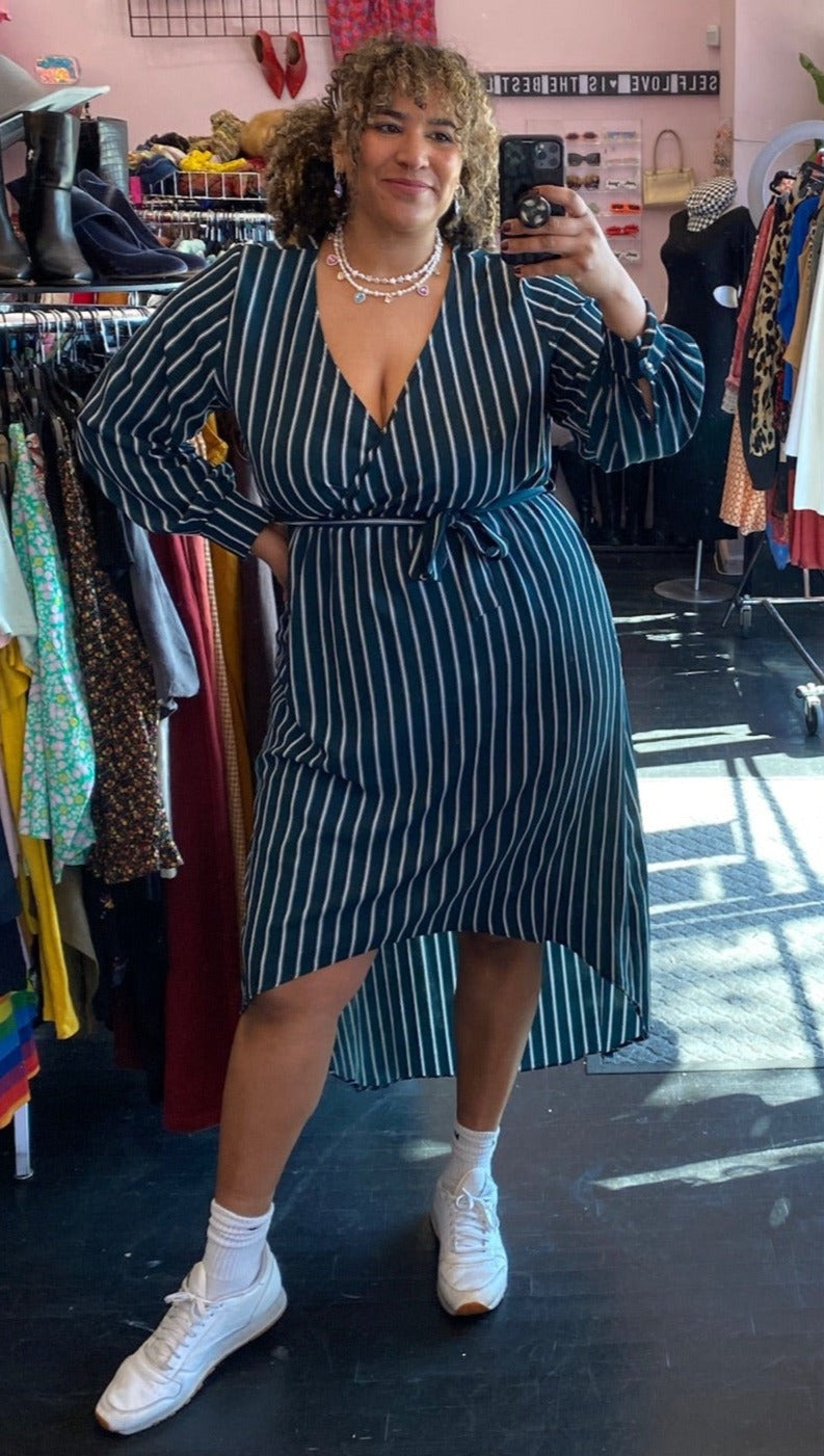 Full-body front view of a size 3X Black Ivy brand dark teal and white vertical striped faux wrap midi dress with high-low hemline styled with white sneakers on a size 16/18 model.