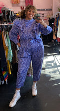 Load image into Gallery viewer, Additional full-body front view of a size 20 Eloquii blue and periwinkle mixed floral print ruffled jumpsuit with puff sleeves and tie belt styled with white sneakers on a size 16/18 model.

