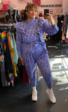 Load image into Gallery viewer, Full-body front view of a size 20 Eloquii blue and periwinkle mixed floral print ruffled jumpsuit with puff sleeves and tie belt styled with white sneakers on a size 16/18 model.
