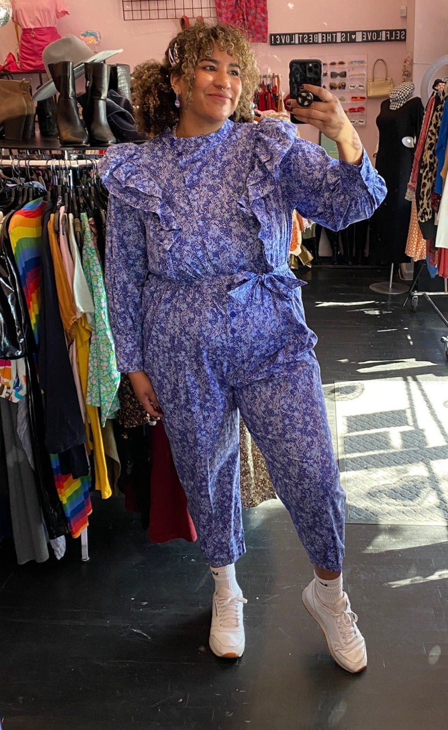 Full-body front view of a size 20 Eloquii blue and periwinkle mixed floral print ruffled jumpsuit with puff sleeves and tie belt styled with white sneakers on a size 16/18 model.