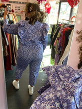 Load image into Gallery viewer, Full-body back view of a size 20 Eloquii blue and periwinkle mixed floral print ruffled jumpsuit with puff sleeves and tie belt styled with white sneakers on a size 16/18 model.

