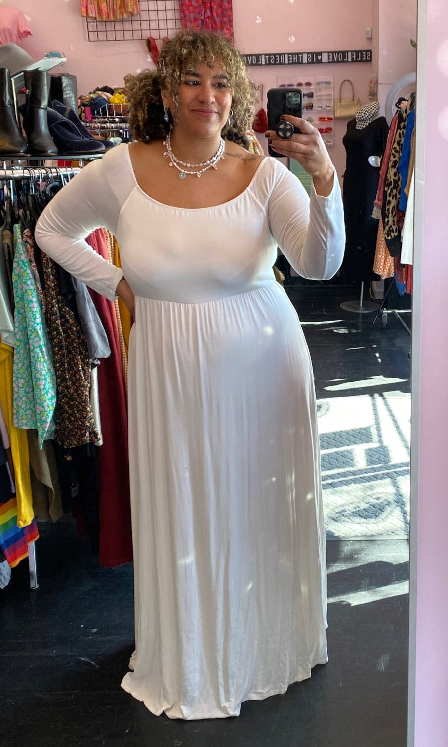 Full-body front view of a size 3X Pink Blush brand off-white floor length maxi dress with scoop neck styled with pearl necklaces on a size 16/18 model. The photo is taken inside in overhead lighting.