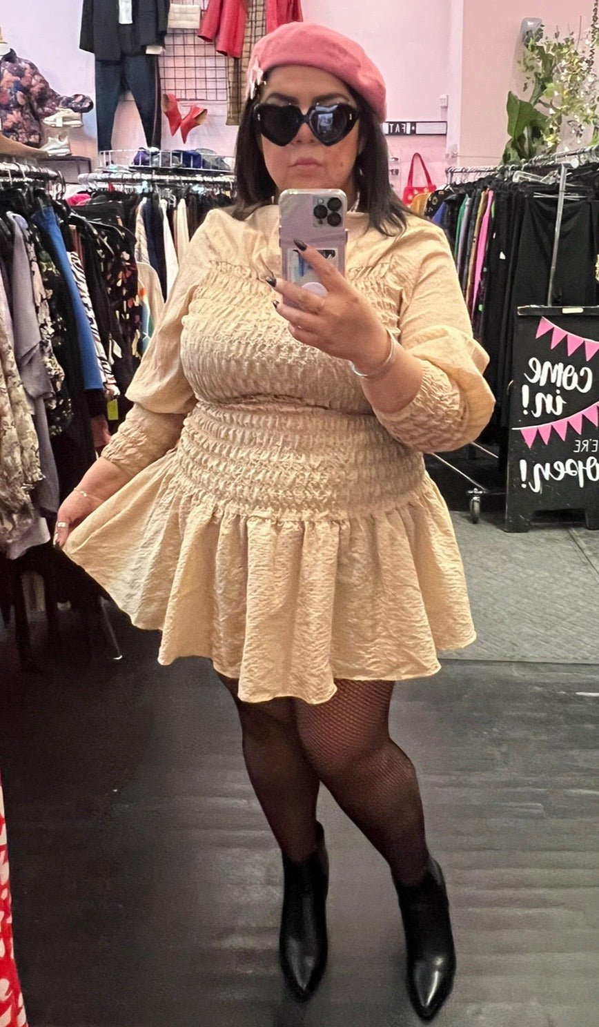 Full-body front view of a size L H&M light cream smocked torso mini dress with puff sleeves and a high neckline styled with black tights and black hoots on a size 14/16 model.