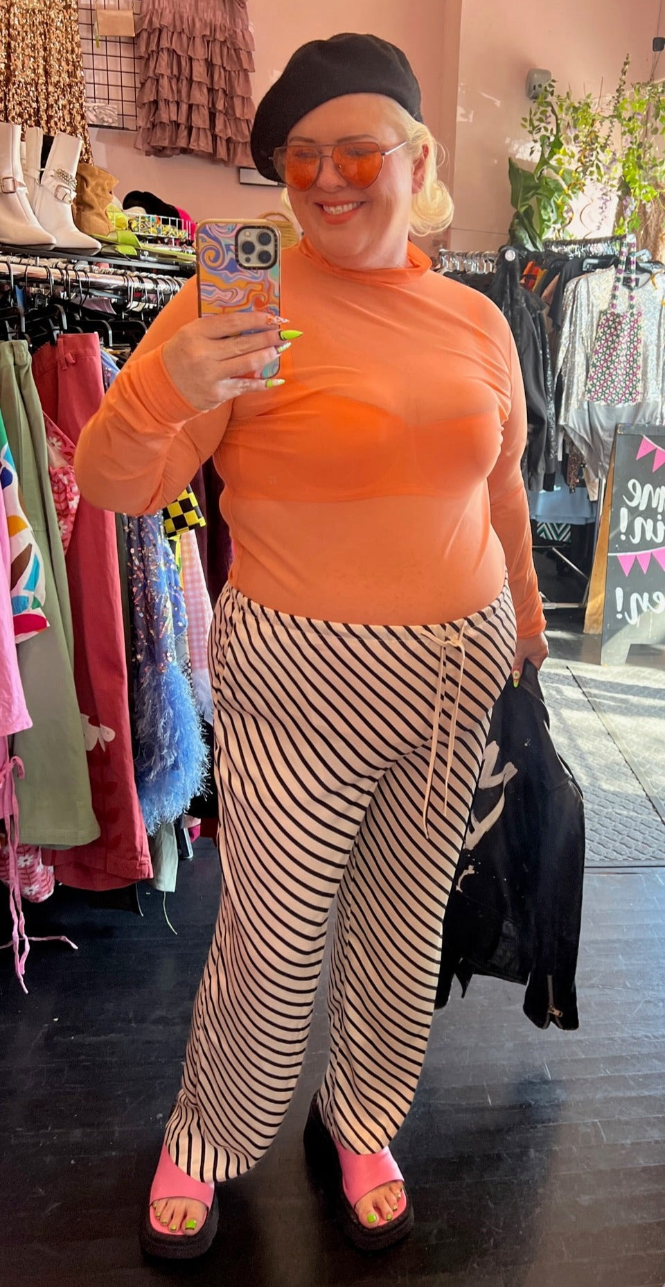 Full-body front view of a pair of size XXL H&M off-white and black diagonally striped silky drawstring pants styled with an orange sheer mesh mockneck top, a black beret, and pink slides on a size 16/18 model.
