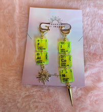 Load image into Gallery viewer, Close up of the green &amp; gold colorway of these Wrapped By Inara &quot;FAT ICON&quot; elemental earrings with a little sun charm and a needle charm on either earring.
