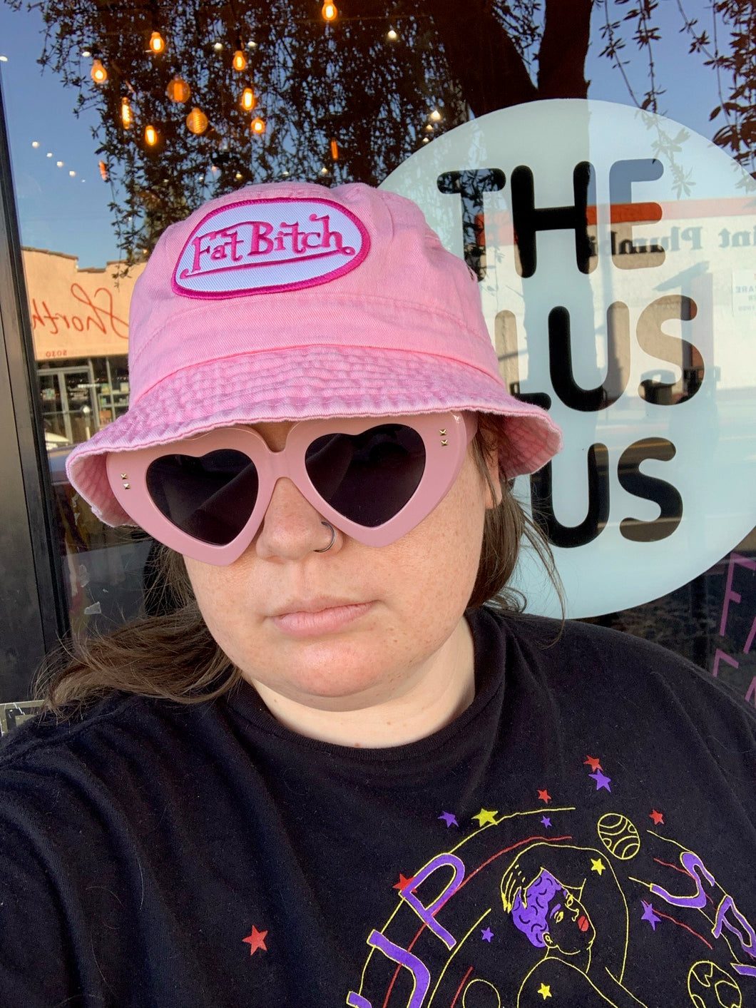 Side-long close up on the pink FAT BITCH bucket hat, a black hat with a white and pink embroidered patch that reads 
