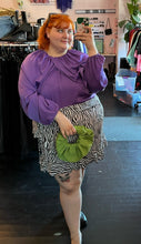 Load image into Gallery viewer, Another full-body front view of a size 28 Eloquii vibrant purple draped and gathered blouse with balloon sleeves styled with a zebra ruffled skirt and a green handband on a size 22/24 model.
