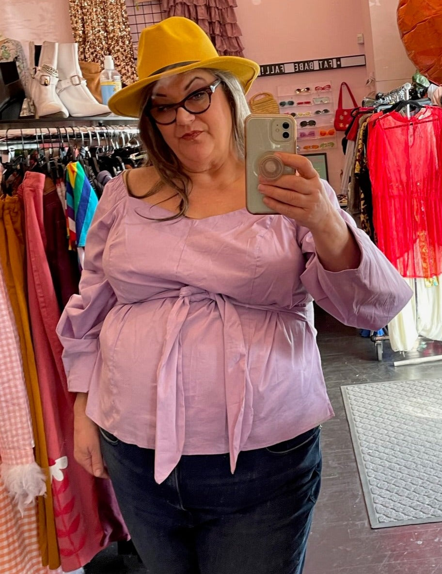 Front view of a size 24 Eloquii lavender square neck long sleeve blouse with tie detail styled with a yellow hat and dark wash jeans on a size 20/22 model.