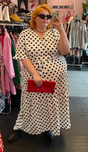 Load image into Gallery viewer, Full-body front view of a size 4X Rebdolls white and black polka dot ruffle-tiered maxi dress with ruffle sleeves and tie belt styled with leather boots, black sunglasses, and a red clutch on a size 22/24 model.
