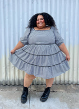 Load image into Gallery viewer, Full-body front view showing the flounce of the skirt of a size 28 Simply Be brand black and white gingham plaid mini-print tiered prairie dress with black trim and puff sleeves styled with black combat boots on a size 24/26 model. The photo is taken outside in natural lighting.
