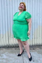 Load image into Gallery viewer, Full-body front view of a size 26 Simply Be brand vibrant kelly green mini dress with all-over swiss dots, ruffle trim and hem, puff sleeves, and a smocked waist styled with black pumps on a size 22/24 model.

