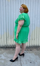 Load image into Gallery viewer, Full-body side view of a size 26 Simply Be brand vibrant kelly green mini dress with all-over swiss dots, ruffle trim and hem, puff sleeves, and a smocked waist styled with black pumps on a size 22/24 model.
