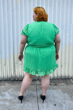 Load image into Gallery viewer, Full-body back view of a size 26 Simply Be brand vibrant kelly green mini dress with all-over swiss dots, ruffle trim and hem, puff sleeves, and a smocked waist styled with black pumps on a size 22/24 model.
