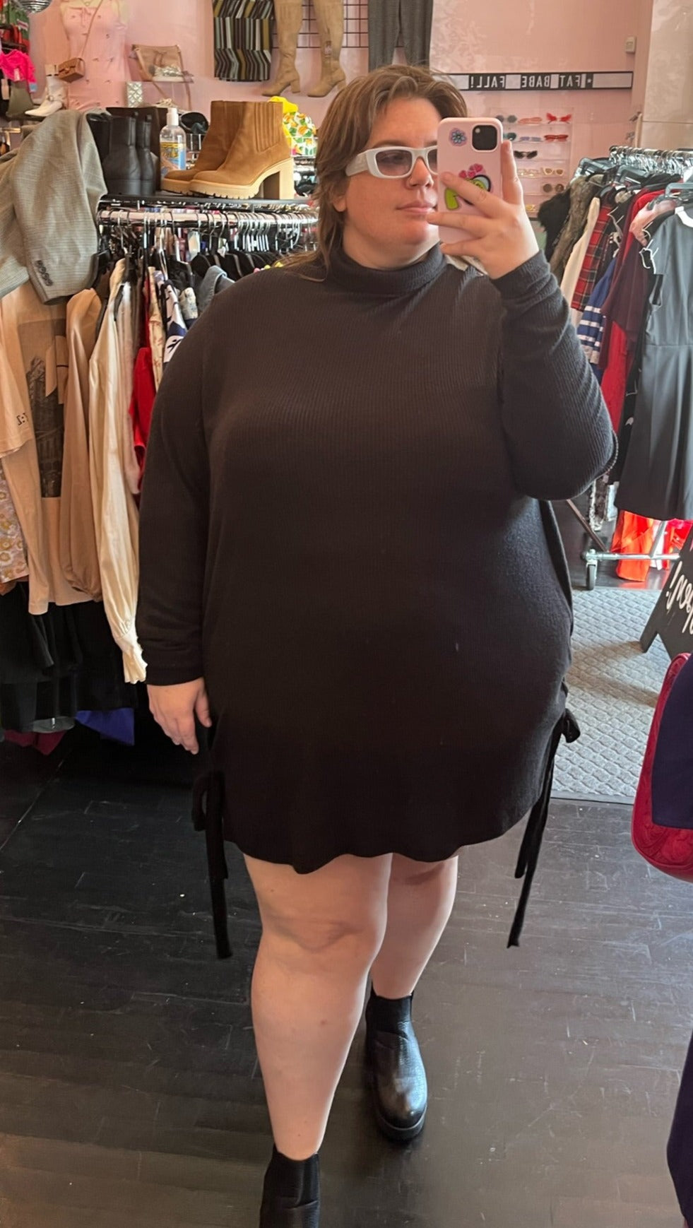 Full-body front view of a size 3X Zelie for She black ribbed turtleneck mini dress with adjustable side details styled with black boots on a size 24 model.
