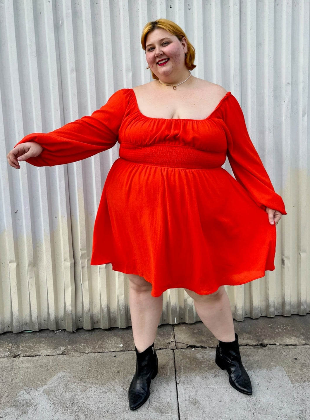 Full-body front view showing off the flounce of a size 2X WRAY red-orange scoop neck mini dress with smocked waist and puff sleeve with elastic cuff styled with black boots on a size 22/24 model.