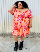 Load image into Gallery viewer, Full-body front view of a size 4X BP peach, pink, and orange floral off-shoulder mini dress with subtle high-low hemline styled with black combat boots on a size 24/26 model.
