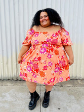 Load image into Gallery viewer, Full-body front view showing off the stretch of the skirt of a size 4X BP peach, pink, and orange floral off-shoulder mini dress with subtle high-low hemline styled with black combat boots on a size 24/26 model.
