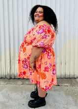 Load image into Gallery viewer, Full-body side view of a size 4X BP peach, pink, and orange floral off-shoulder mini dress with subtle high-low hemline styled with black combat boots on a size 24/26 model.
