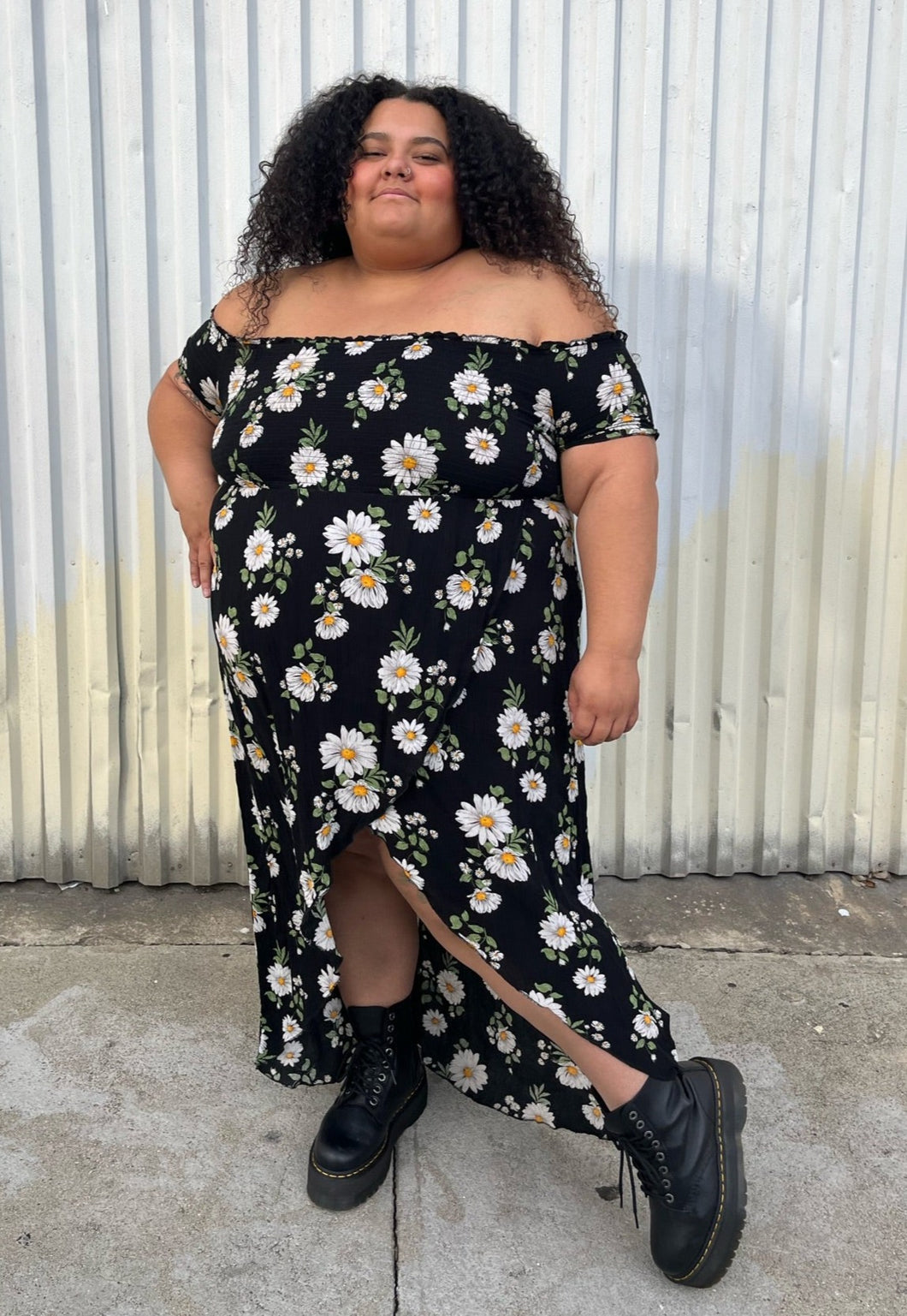 Full-body front view of a size 2X Forever 21 black, white, and yellow daisy pattern smocked bust off-shoulder maxi dress with high-low tulip hem styled with black combat boots on a size 24/26 model. The photo is taken outside in natural lighting.