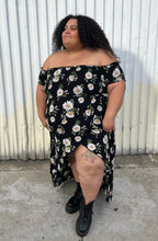 Load image into Gallery viewer, Additional full-body front view showing off the tulip hem slit of a size 2X Forever 21 black, white, and yellow daisy pattern smocked bust off-shoulder maxi dress with high-low tulip hem styled with black combat boots on a size 24/26 model. The photo is taken outside in natural lighting.
