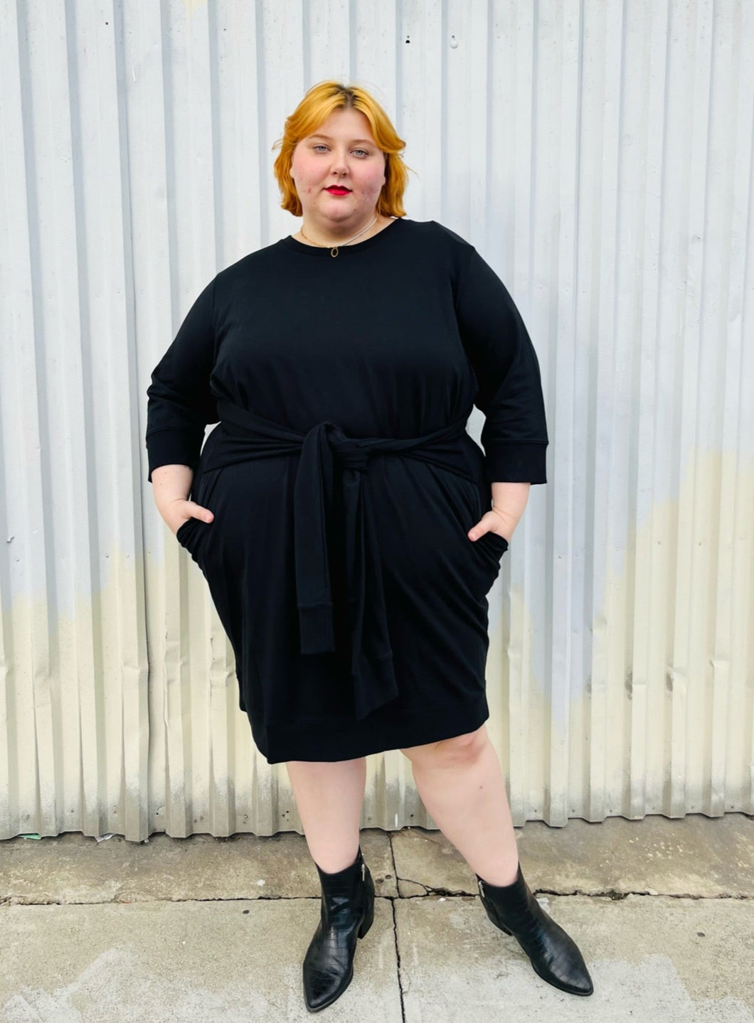 Full-body front view of a size XL (size 24/26) Universal Standard black sweatshirt-style crew neck dress with waist tie detail styled with black pointy boots on a size 22/24 model. The photo is taken outside in natural lighting.