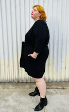 Load image into Gallery viewer, Full-body side view of a size XL (size 24/26) Universal Standard black sweatshirt-style crew neck dress with waist tie detail styled with black pointy boots on a size 22/24 model. The photo is taken outside in natural lighting.
