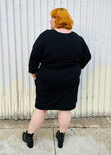 Load image into Gallery viewer, Full-body back view of a size XL (size 24/26) Universal Standard black sweatshirt-style crew neck dress with waist tie detail styled with black pointy boots on a size 22/24 model. The photo is taken outside in natural lighting.
