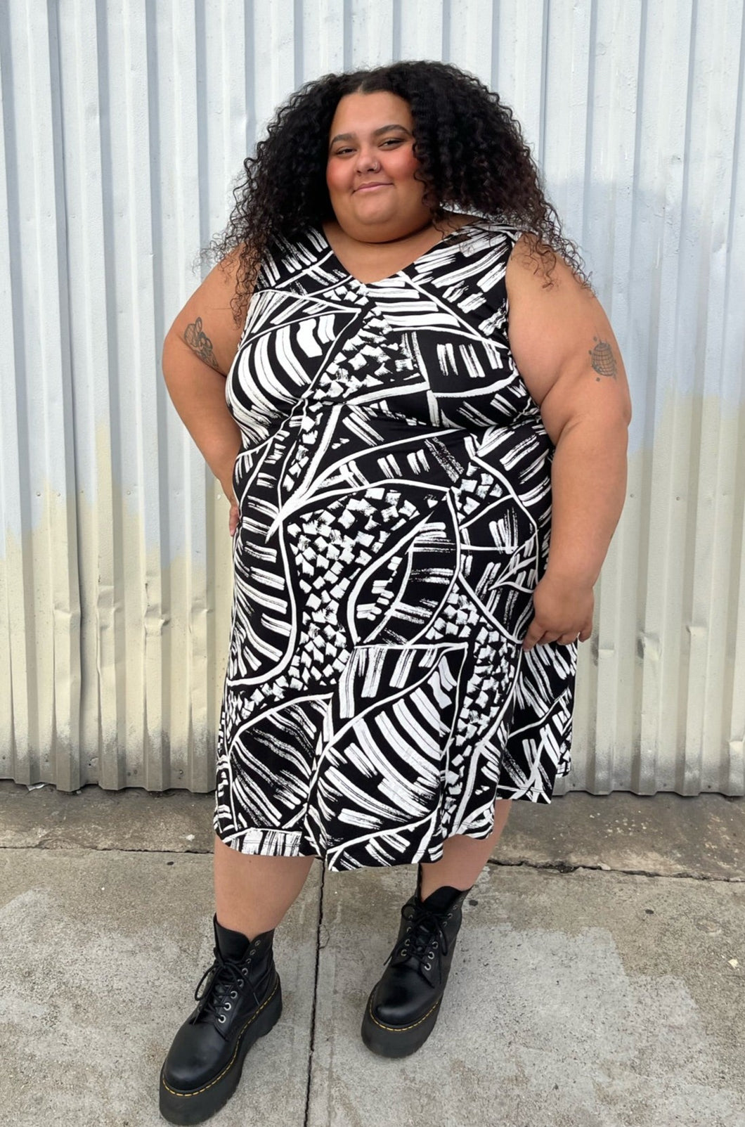 Full-body front view of a size 3X Nic+Zoe black and white abstract geometric print tank midi dress styled with black combat boots on a size 24/26 model. The photo is taken outside in natural lighting.