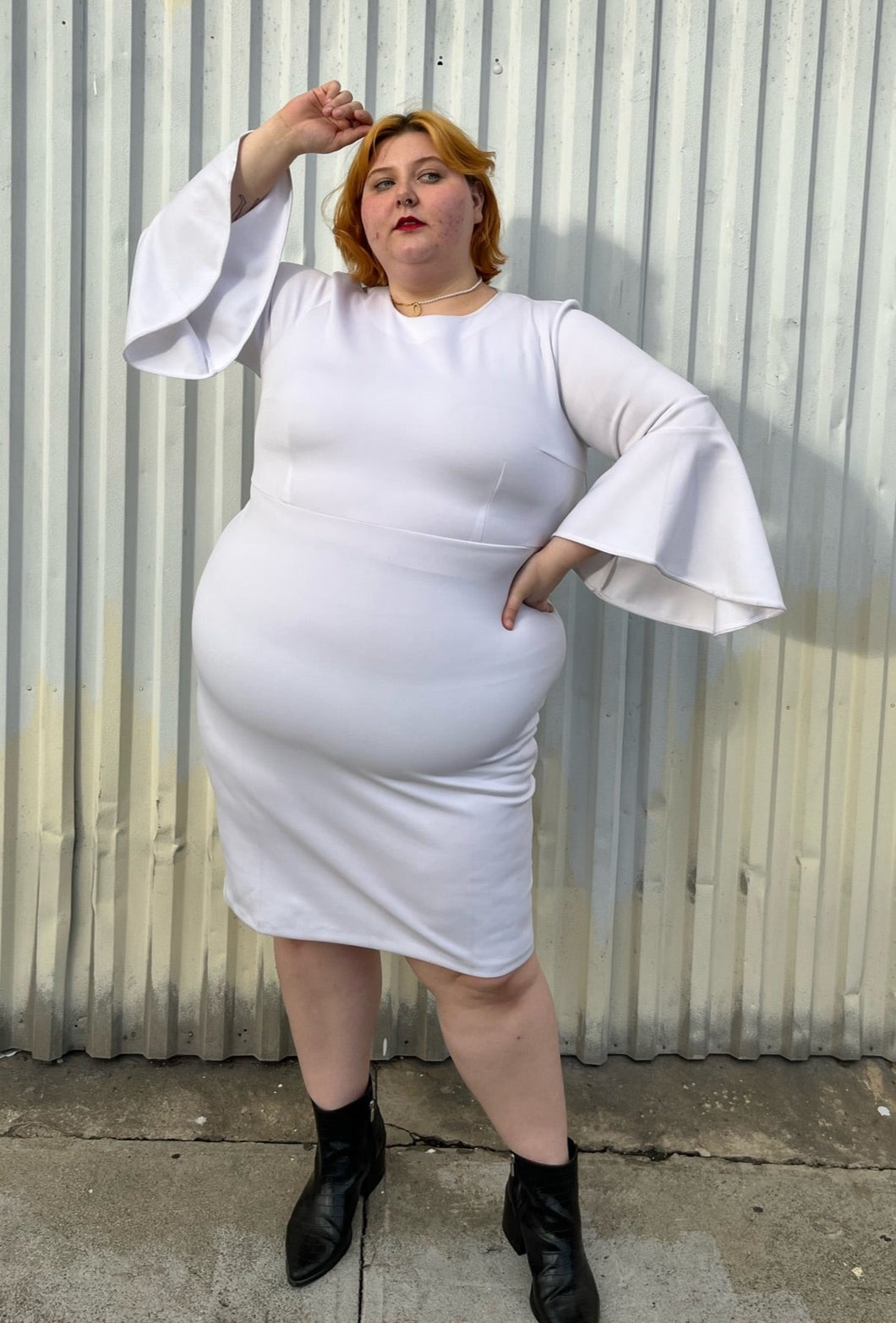Full-body front view showing off the length and flow of the bell sleeves of a size 24 Eloquii white shift dress with long bell sleeves styled with black pointy boots on a size 22/24 model. The photo is taken outside in natural lighting.