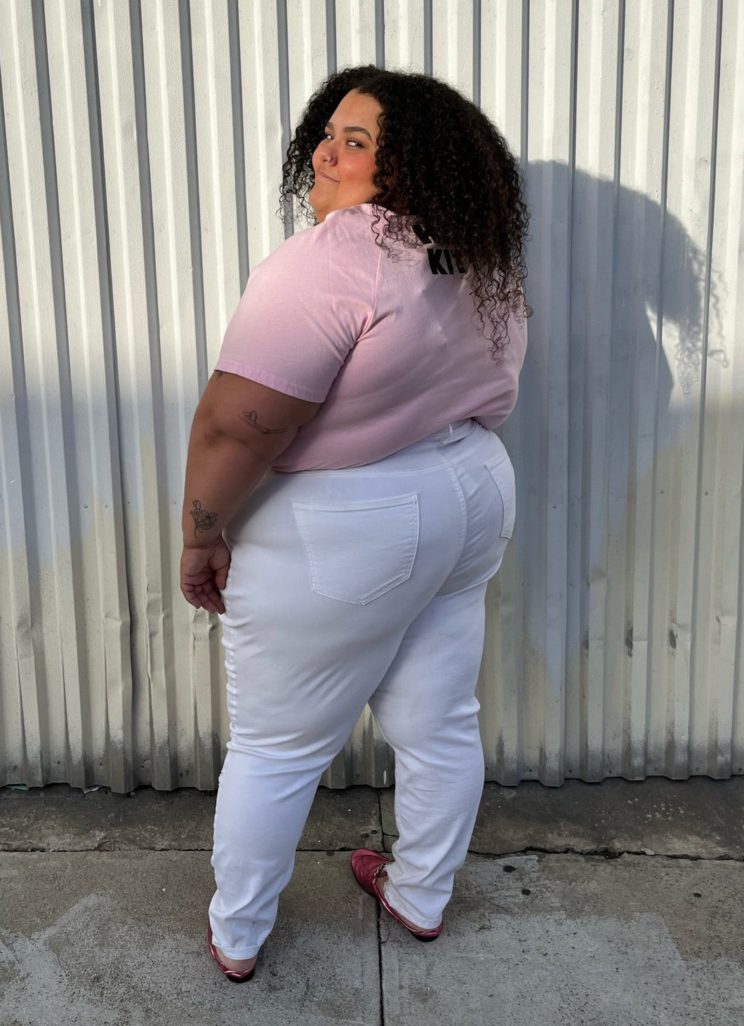 Full-body back view of a pair of size 24 Fashion to Figure slight high-waisted white distressed denim with the distressing focused at the knee and shins styled with a pink tee and pink loafers on a size 24/26 model.