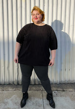Load image into Gallery viewer, Full-body front view of a size 24 ASOS black plisse pleated tee styled over black and white windowpane pattern pants with black point boots on a size 22/24 model. The photo is taken outside in natural lighting.
