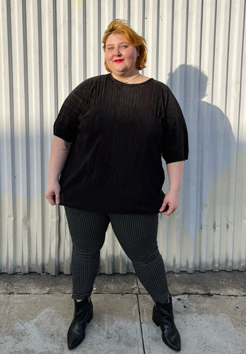 Full-body front view of a size 24 ASOS black plisse pleated tee styled over black and white windowpane pattern pants with black point boots on a size 22/24 model. The photo is taken outside in natural lighting.