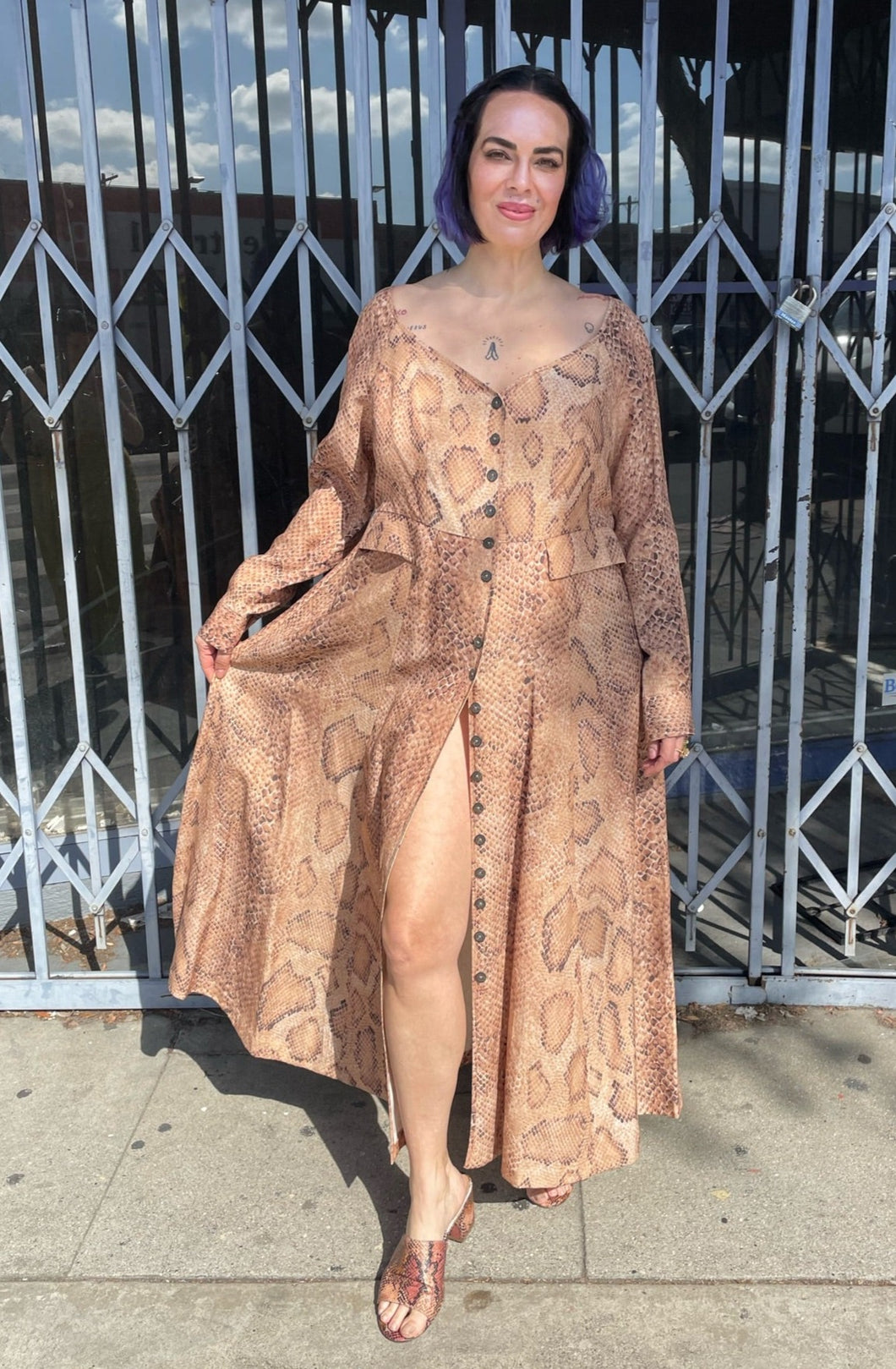 Full-body front view showing off the slit of a size 12 Mara Hoffman tan, brown, and white snake print button-up maxi dress with hip pocket details styled with snake print heels on a size 10 model. The photo is taken outside in natural lighting.