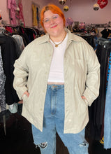 Load image into Gallery viewer, Front view of a size 2/3X Public Desire brand off-white faux leather longline snap button-up top styled open over a white cropped tee and lightwash denim on a size 22/24 model.
