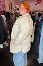 Load image into Gallery viewer, Side view of a size 2/3X Public Desire brand off-white faux leather longline snap button-up top styled open over a white cropped tee and lightwash denim on a size 22/24 model.

